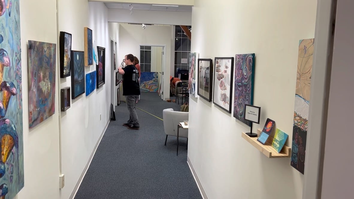Gallery hallway lined with diverse paintings opening into a back exhibition space