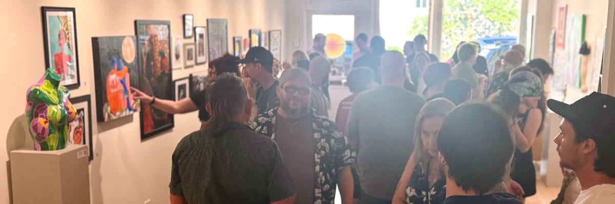 A crowded gallery opening: visitors fill a long, sunlit room lined with framed work, one pointing at a painting beside a flower-patterned torso sculpture.