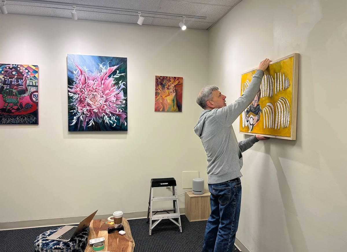 Gallerist hanging a framed artwork on a white wall beside an installed exhibition