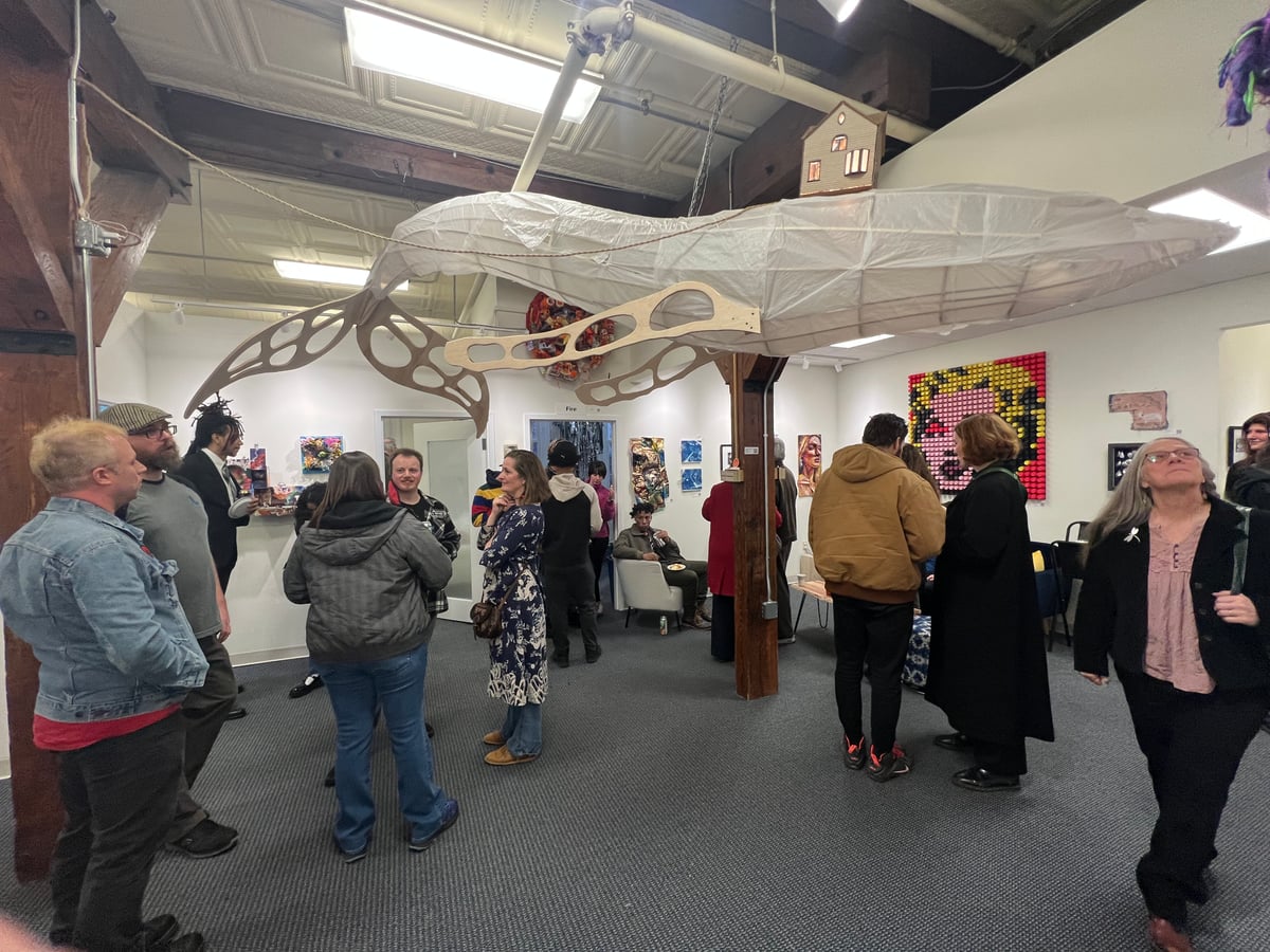 Gallery opening with visitors gathered beneath a hanging paper-and-wood butterfly sculpture